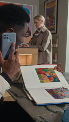 Vertical of young Black man sitting at table in modern art gallery looking at museum catalog with abstract paintings and talking on phone with Caucasian female colleague working in background