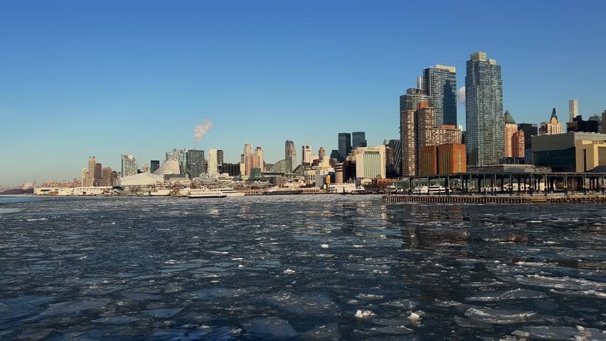 Wide view shows Manhattan skyline, piers, ferry across icy Hudson River on sunny winter day. Ideal establishing shot for urban winter, NYC, USA