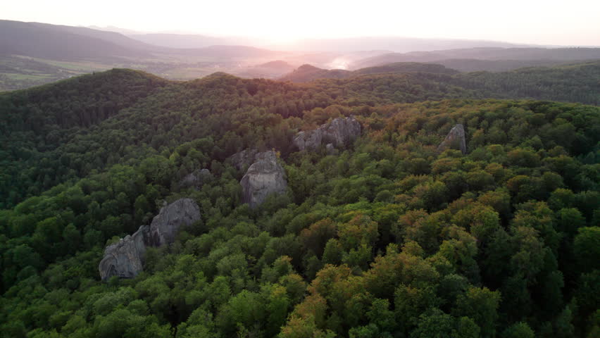 Aerial view of rock formation bathed in warm sunlight among lush green forest and rolling hills. Dovbush Rocks, Carpathian mountains, Ukraine.