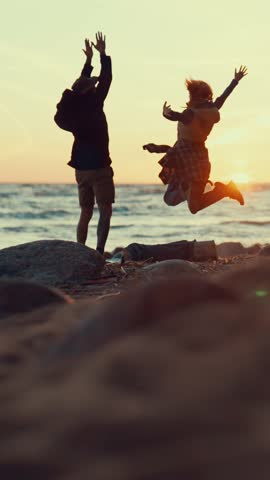 Joyful man and woman admiring sunset on sea shore and jumping up, fun and joy. People in nature, be happy and carefree, summer vacation and holidays, vertical shot, slow motion, active pastime in park