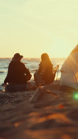 Cozy summer evening on seashore, loving couple wrapped in blanket admiring sunset. Touristic camp on shore, back view of tired backpackers resting in nature, local tourism and hiking, vertical video