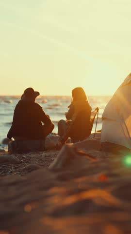 Cozy summer evening on seashore, loving couple wrapped in blanket admiring sunset. Touristic camp on shore, back view of tired backpackers resting in nature, local tourism and hiking, vertical video