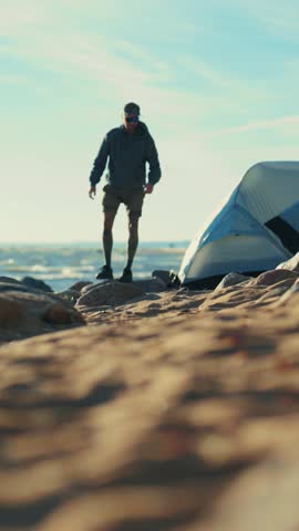 Summer trip to sea, man traveler pitching tent on shore of sea or lake in sunny day, vertical shot. Local tourism and hiking in beautiful natural park, exploring shoreline, discovery and adventure