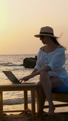 Young woman in sunglasses and sun hat, uses laptop, sitting on the beach by the sea, at sunset or sunrise. freelancer, remote work. work and vacation at sea