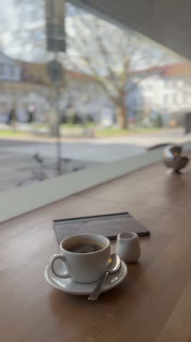 Cup of black coffee and milk on a wooden table by a large window with a soft view of the city street. Cozy indoor cafe scene. Vertical video.