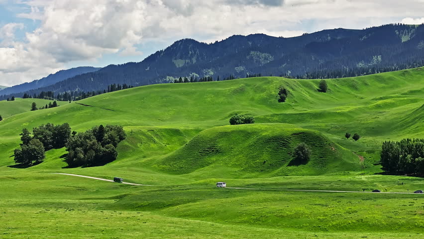 Picturesque view of vibrant green rolling hills with a road and distant mountains under cloudy sky. Beautiful grassland natural landscape in Nalati Grassland, Xinjiang, China.