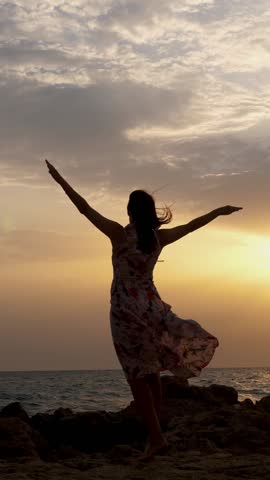 Dark silhouette of a young woman standing by the sea, at sunrise. the wind blows her dress