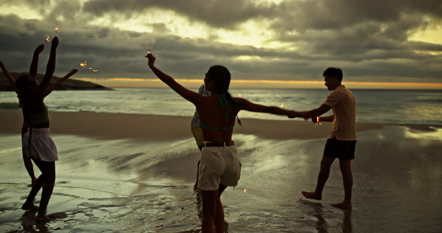 Happy, friends and dancing with sparkler on beach for ocean party, travel reunion and bonding together. People, fireworks and rhythm movement, New years celebration and sea sunset on freedom vacation