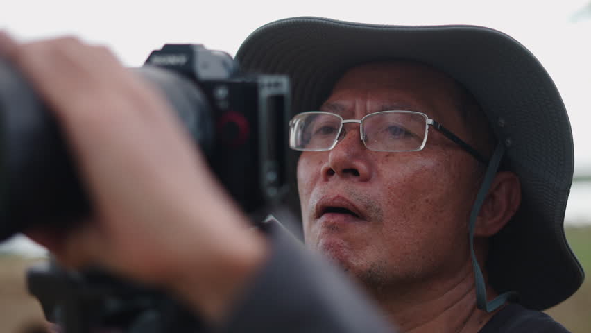 closeup of asian photographer face with glasses and hat adjusting the focus of his camera
