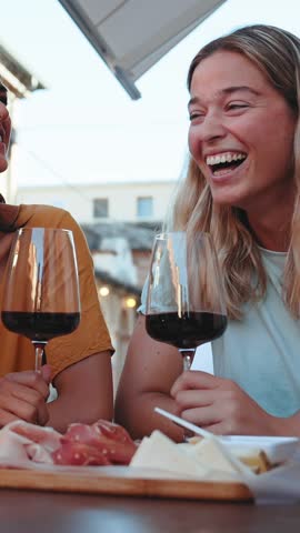 Vertical. Three happy female friends are toasting with glasses of red wine while sitting at a table outside a restaurant in a picturesque Italian town, enjoying a selection of cheese and cured meats