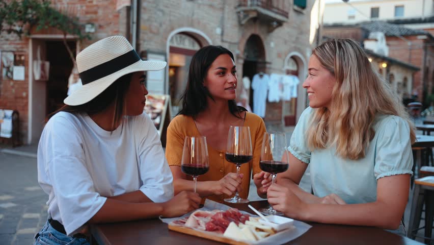 Three happy female friends are toasting with glasses of red wine while sitting at a table outside a restaurant in a picturesque Italian town, enjoying a selection of cheese and cured meats
