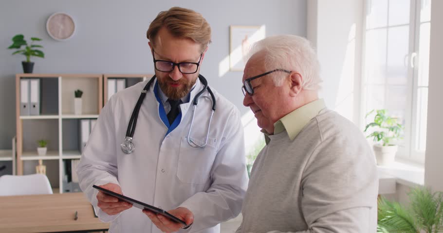 Elderly male patient consulting with a doctor or nurse using a tablet pc during a hospital or clinic visit. Modern healthcare, medical care, and digital online communication in medicine.
