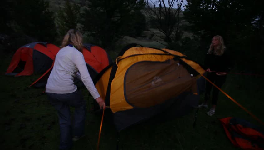 A medium shot of two girls pitching a tent in wilderness at night.