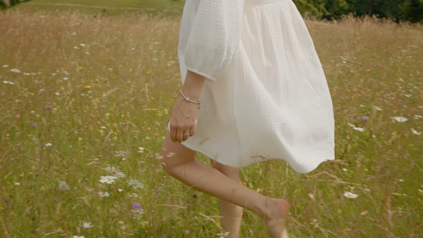 Pregnant woman in a flowing white dress walks gracefully through vibrant flowering grass. Surrounded by mountains and fir trees, she embraces nature and relaxation.