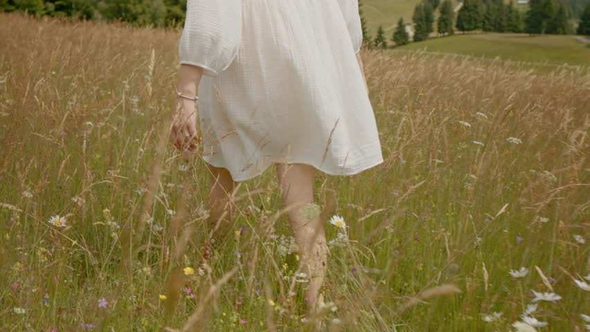 Pregnant woman in a flowing white dress walks gracefully through vibrant flowering grass. Surrounded by mountains and fir trees, she embraces nature and relaxation.