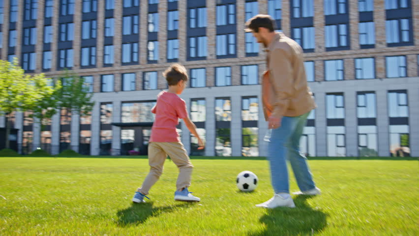 Single father son playing football on grass near house. Happy family kid parent enjoying time together. Positive man boy having fun with ball in summer park place. Daddy child relationships concept 