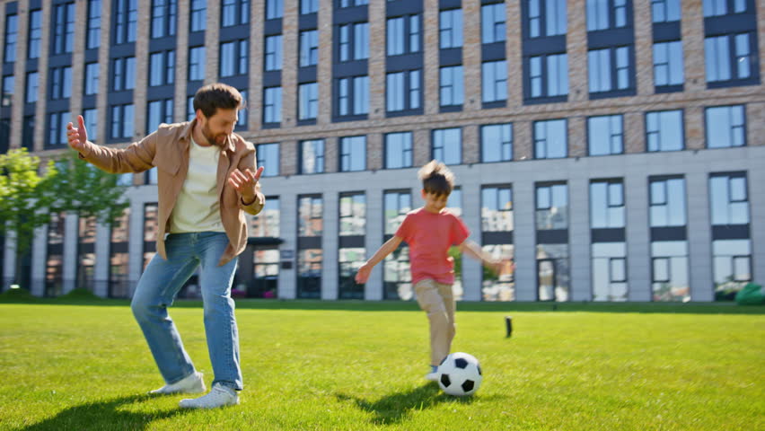 Single father son playing football on grass near house. Happy family kid parent enjoying time together. Positive man boy having fun with ball in summer park place. Daddy child relationships concept 