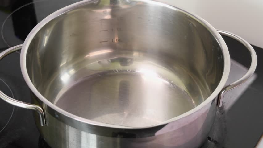 Clear water pours into shiny stainless steel pot on black stove in bright kitchen, preparing for cooking or cleaning process.