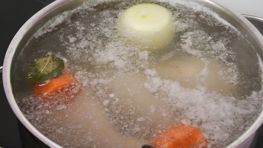 Foamy meat broth with onion, carrot and bay leaf boiling in large pot during cooking process, fat spots floating on surface