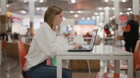 Young woman working on laptop in mall food court, seated at table in modern interior, typing and focused on screen with vibrant background of blurred people and bright lights - Powered by Shutterstock - Get 15% off with code: PIKWIZARD15