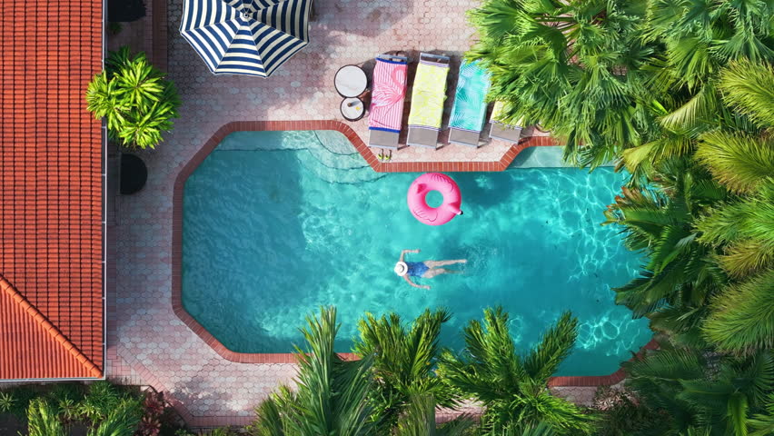 Overhead slow motion video of woman swimming smoothly in tropical backyard pool, captured in bright sunlight with a pink flamingo float nearby, striped umbrella, colorful loungers, and lush palm trees