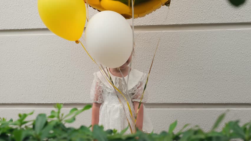 A young girl in a white dress holds a large golden balloon while surrounded by colorful balloons outdoors.