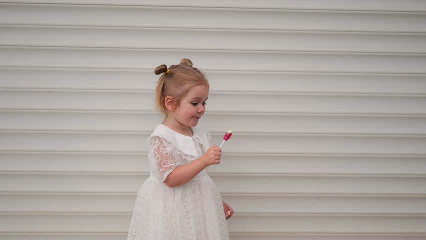 A girl wearing a white dress and black boots happily licks a lollipop outside on a bright day.