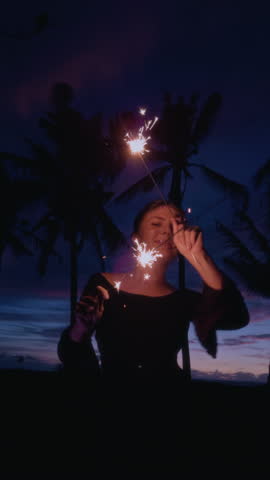 Woman holding sparkler. Christmas lights at the beach. Girl celebrate new year eve in tropical palm tree. Female and Bengal fire. Slow motion vertical video. Blue hour and coconut tree