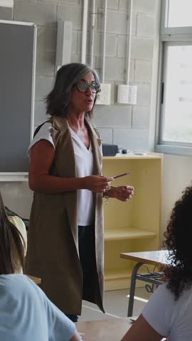 Vertical HD video of senior female teacher explaining lesson to attentive multi-ethnic students in classroom at high school. Education lifestyle concept