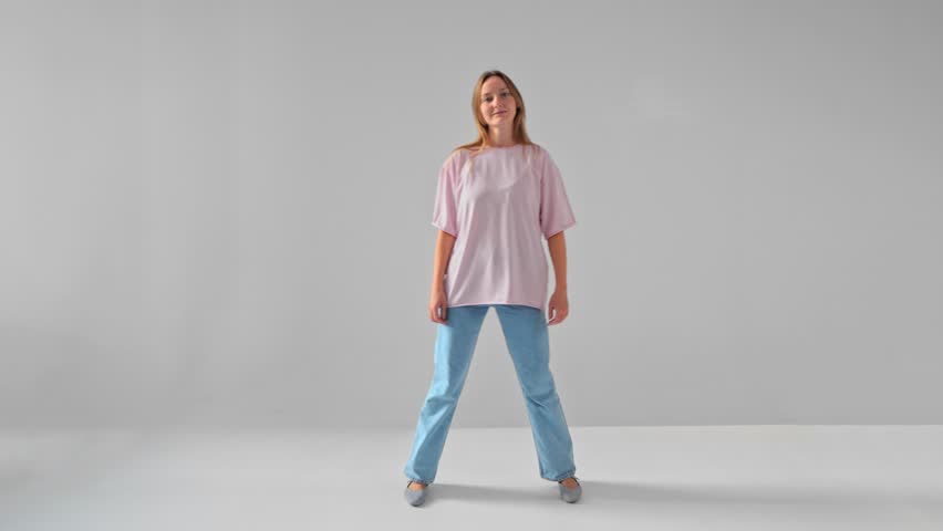 Woman is stepping sideway to the right standing in studio against white background. Isolated full length view of young adult Caucasian lady wearing jeans and T-shirt moving and looking at camera