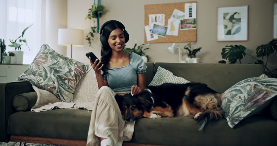 Phone, music and happy woman in home with dog for dance, streaming hip hop and rock online. Headphones, pet and listening to audio, radio sound and Indian girl relax on living room sofa with mobile