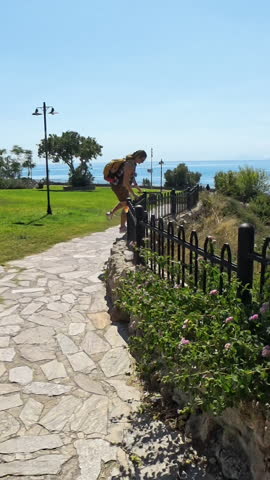 Vertical video. A Man and Woman walks along the edge of a stone wall next to a steep drop, with green grass and blue skies behind him. The scene is filmed using a wide-angle lens, capturing the surrou