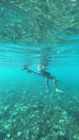 Young surfer sit on his surf board in transparent tropical water of Indian Ocean. Summer dream vacations. Low angel wide short. Underwater vertical video. Slow motion. Bali. Indonesia