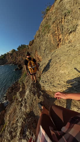 Vertical video. Following a Hiker Along a Narrow Cliff Path with Jagged Rocks and a View of the Sea Glistening in the Sun.
