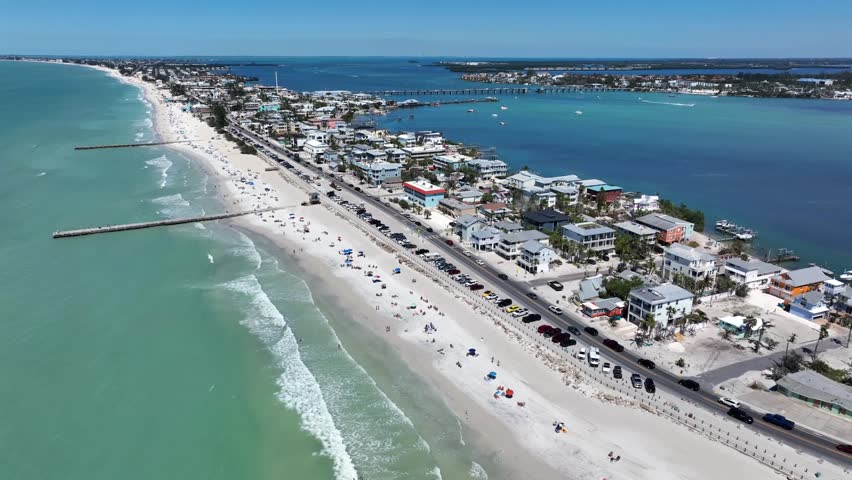 Hotels and Luxury Houses with traffic on main street of Bradenton beach. Jetty with turquoise golf of mexico and blue bay on other side. Aerial backwards wide shot.