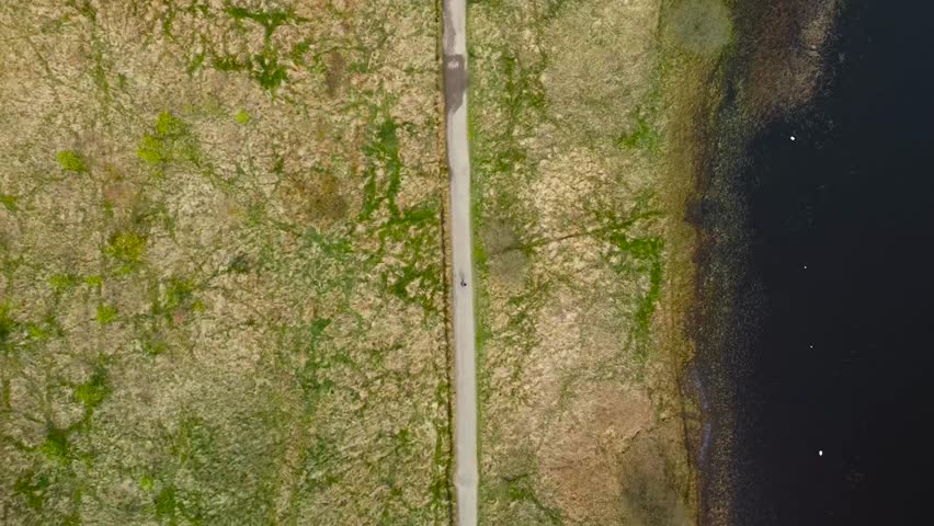 Top down aerial drone footage view of a person walking on a desolate rural paved road in Scotland british green moss covered nature besides a large lake that is dark colored during beautiful day time.