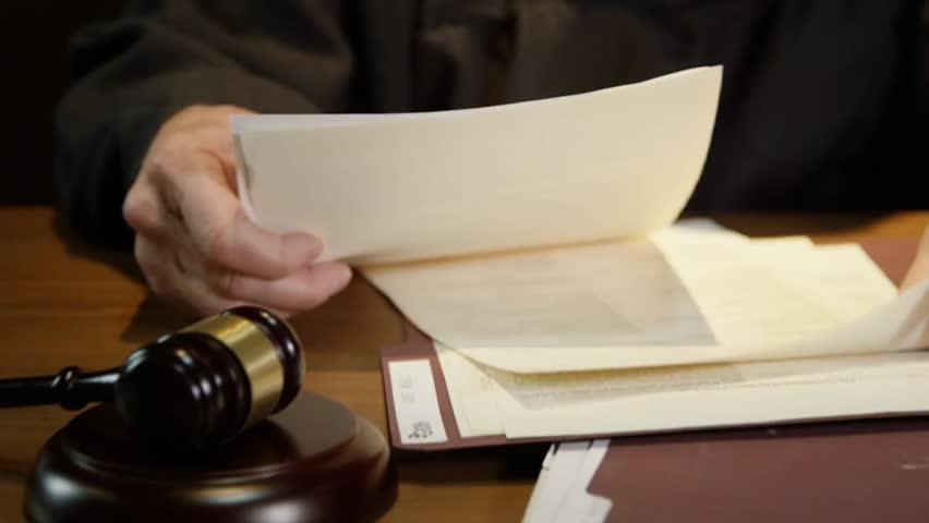 A judge reviewing legal documents in court, carefully examining the paperwork before making a decision. The focus is on the judge's hand and the documents, highlighting the seriousness of the legal pr