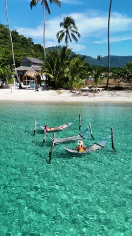 Tropical luxury unfolds with two hammocks gently swaying in the sunlit sea. Surrounded by lush palm trees and soft white sands of Koh Kood Thailand