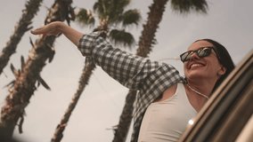 Excited woman enjoys the palm trees of seaside road. A girl leans out of window of rental car on a coastal highway on a sunny day. Hair in the wind. Tropical vacation on the beach of exotic islands - Powered by Shutterstock - Get 15% off with code: PIKWIZARD15
