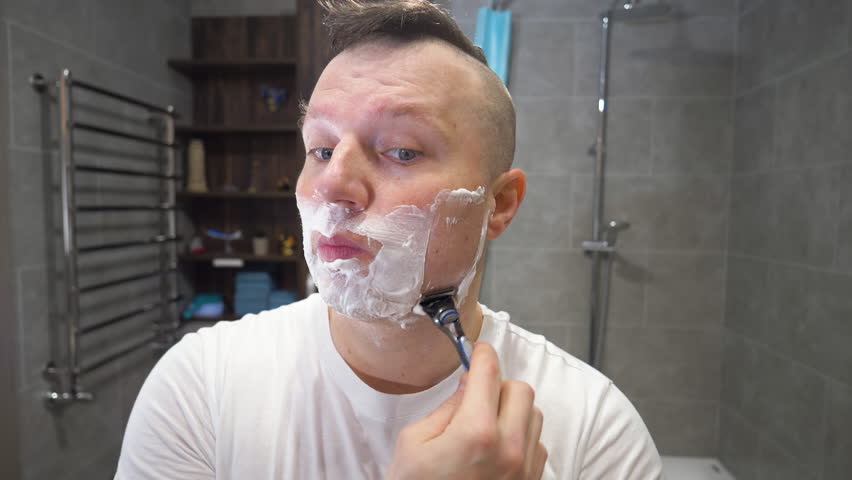 Shave with a razor blade and shaving foam. A young man shaves his beard and stubble with a machine with a blade. slow motion.