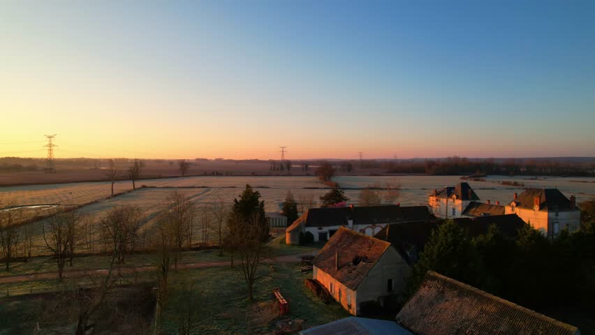 Peaceful aerial view of a traditional French farm bathed in warm sunrise light. Frost-covered fields, rustic buildings, and countryside charm create a serene morning scene.