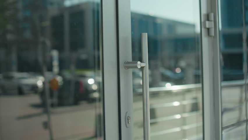 Close up of young lady with handbag opening glass door entering office building, focus on hand gripping metal handle, reflection of woman visible on door, urban professional environment