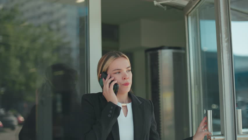 Frustrated woman during phone call lowers phone briefly, continues conversation after work while exiting office building, dressed in formal attire, glass door reflects cityscape