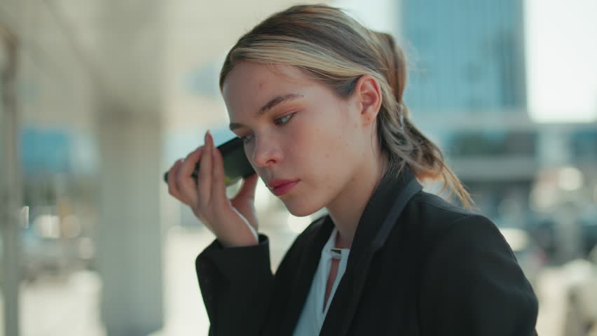 Close up of female lawyer attentively listening to voice note from client while standing outside modern glass building reflecting surroundings