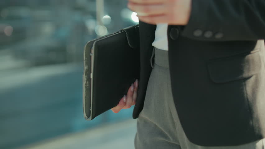 Close up waist view of lady in black blazer and trousers holding tab while walking by building with glass facade reflecting urban blur background in soft sunlight