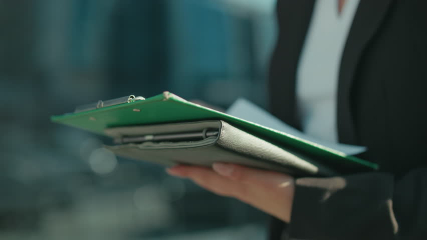 Close up of professional individual holding folder and documents, cross checking details while standing outdoors with glass building in background, focused on reviewing information