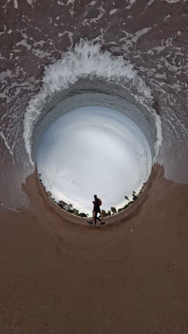Vertical video. A "tiny planet" view captures a man and a woman walking along the curved shoreline. Waves encircle the spherical perspective, emphasizing the expansive beach and ocean
