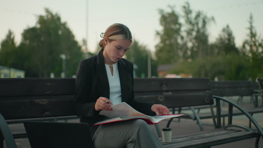 Young girl seated with crossed leg flips through folder in hand with laptop and coffee cup by side on outdoor bench in calm urban setting surrounded by trees and empty benches during daylight