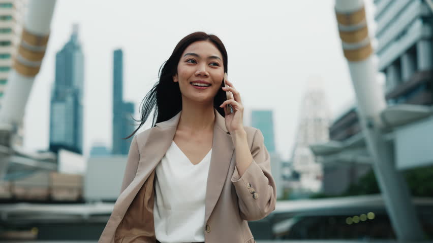 Portrait of joyful Asian lady corporate employee answering phone call in modern city. Thai female happy to talk to business partner on cellphone. Businesswoman conducts conversation