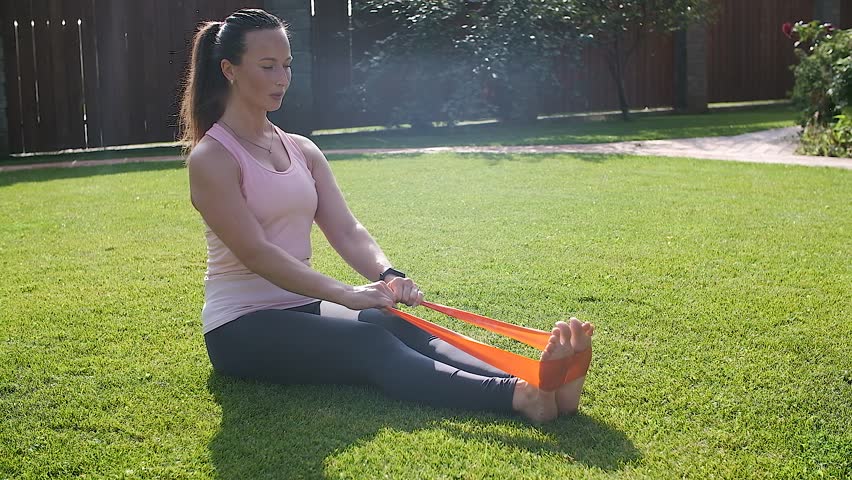 A young woman sitting on the grass doing fitness exercises with an expander strengthening her back muscles.